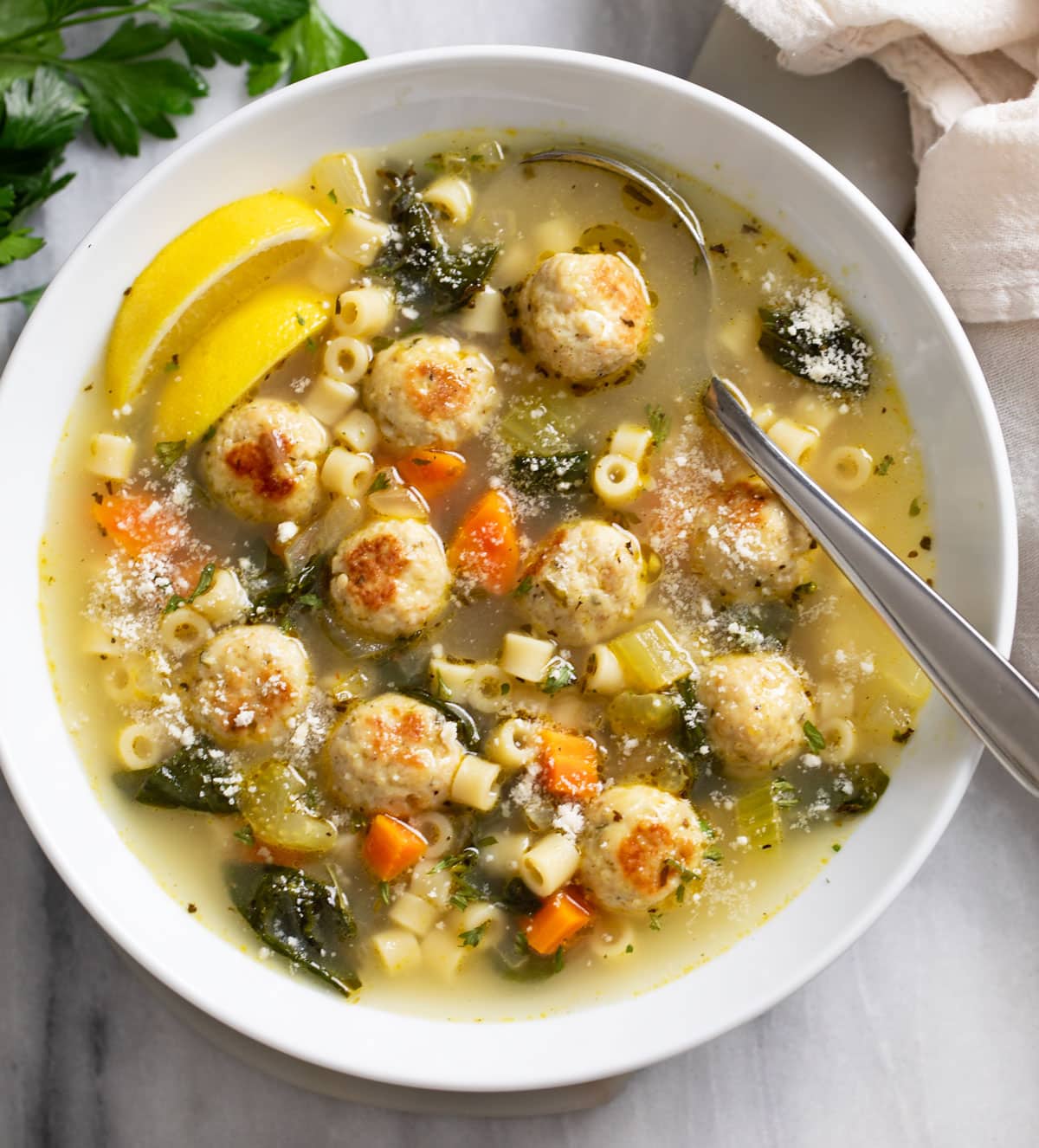 Chicken Meatball Soup in a white serving bowl with lemon wedges and a spoon on the side.