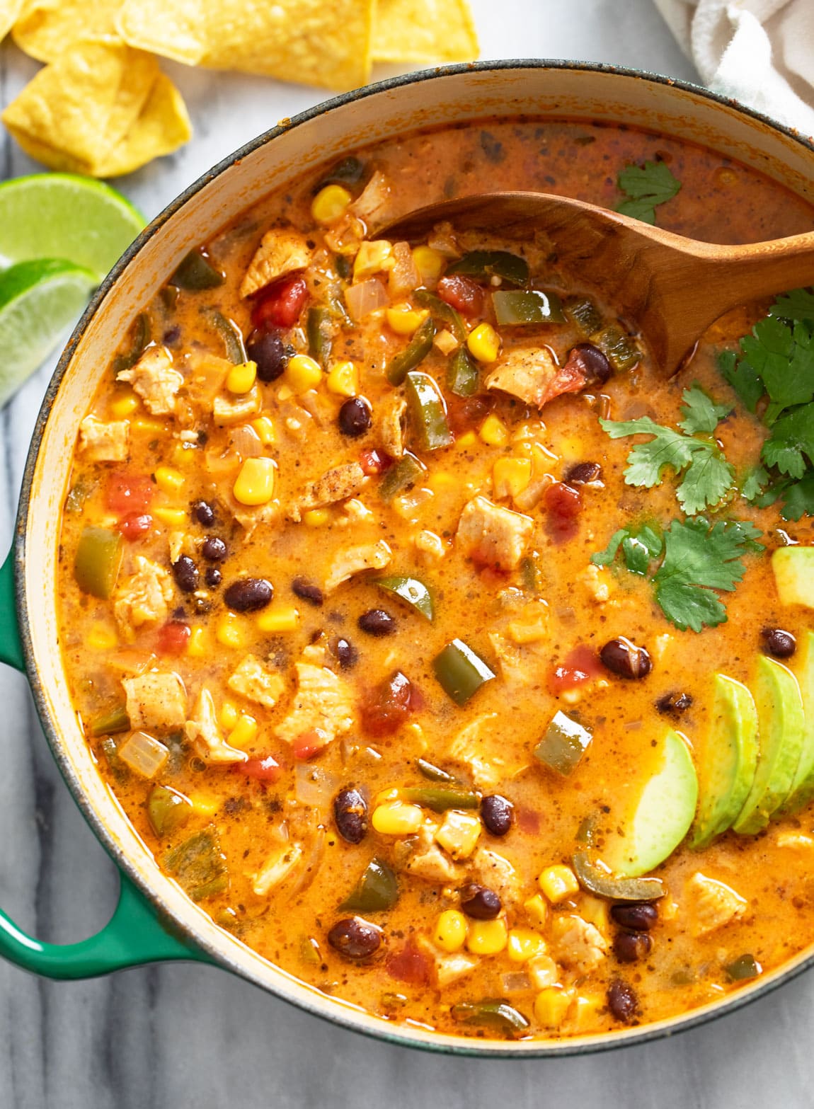 A soup pot filled with Chicken Poblano Soup topped with cilantro and avocado slices with a wooden spoon on the side.