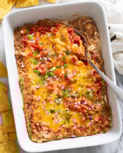 Overhead view of Bean Dip with toppings in a white baking dish with a serving spoon on the side.