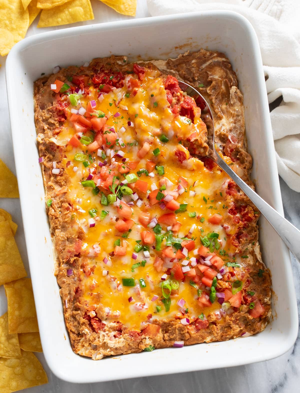Overhead view of Bean Dip with toppings in a white baking dish with a serving spoon on the side.
