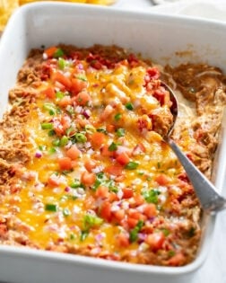 Side view of a white casserole dish filled with Bean Dip, toppings, and a spoon on the side.