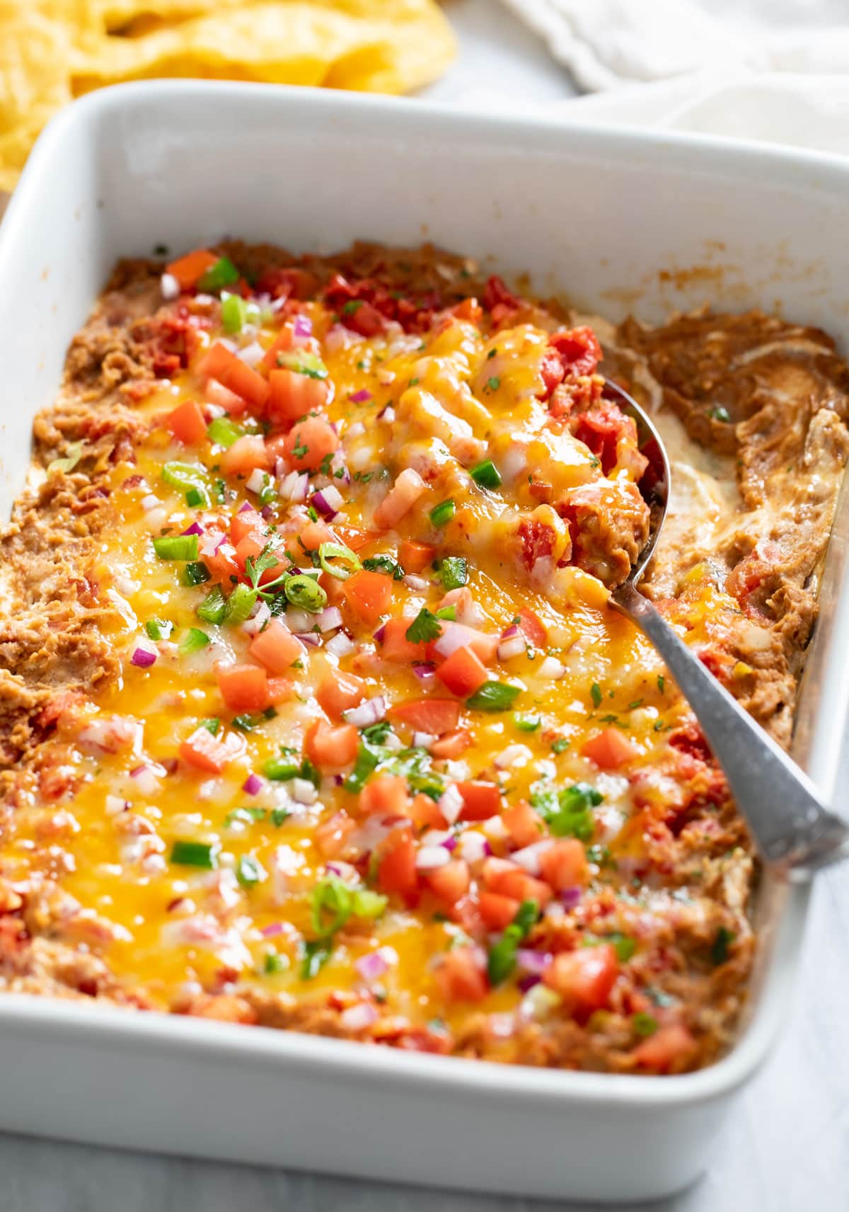 Side view of a white casserole dish filled with Bean Dip, toppings, and a spoon on the side.