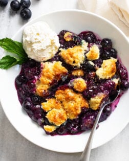 Blueberry Cobbler in a white bowl with a scoop of vanilla ice cream and a sprig of mint on the side.