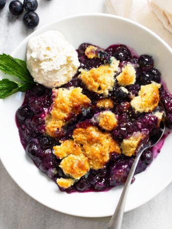 Blueberry Cobbler in a white bowl with a scoop of vanilla ice cream and a sprig of mint on the side.