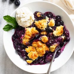 Blueberry Cobbler in a white bowl with a scoop of vanilla ice cream and a spoon on the side.