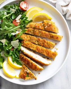 Slices of Chicken Milanese on a white plate with an arugula salad, shaved Parmesan, and lemon wedges.