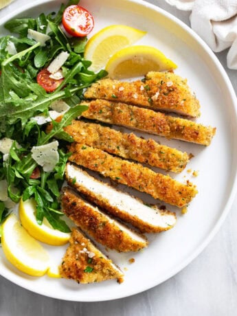Slices of Chicken Milanese on a white plate with an arugula salad, shaved Parmesan, and lemon wedges.