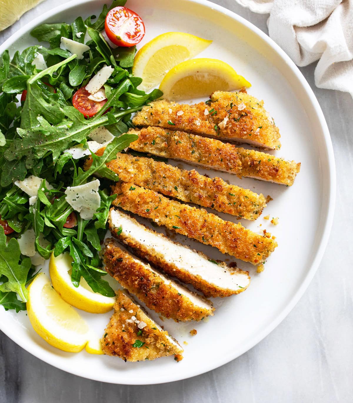 Slices of Chicken Milanese on a white plate with an arugula salad, shaved Parmesan, and lemon wedges.