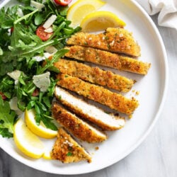 Crispy Chicken Milanese on a plate with lemon slices an arugula salad on the side.