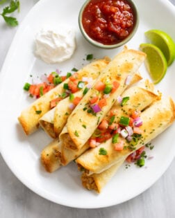 Chicken Taquitos on a white serving platter with salsa, sour cream, lime slices, and toppings.