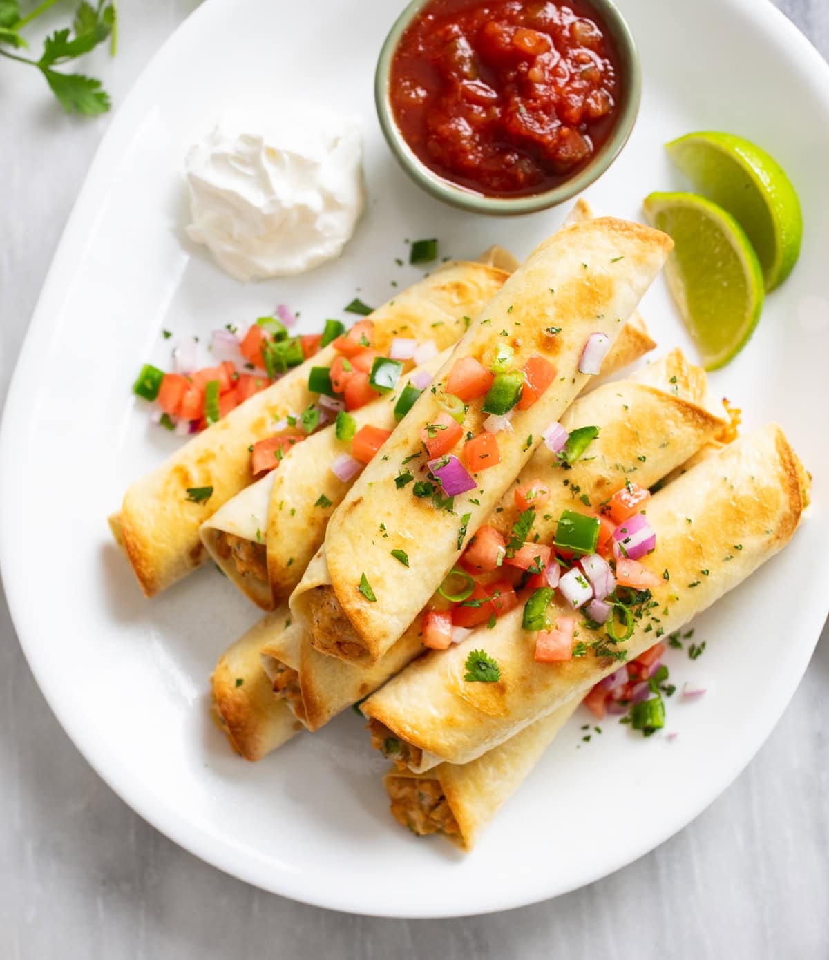 Chicken Taquitos on a white serving platter with salsa, sour cream, lime slices, and toppings.