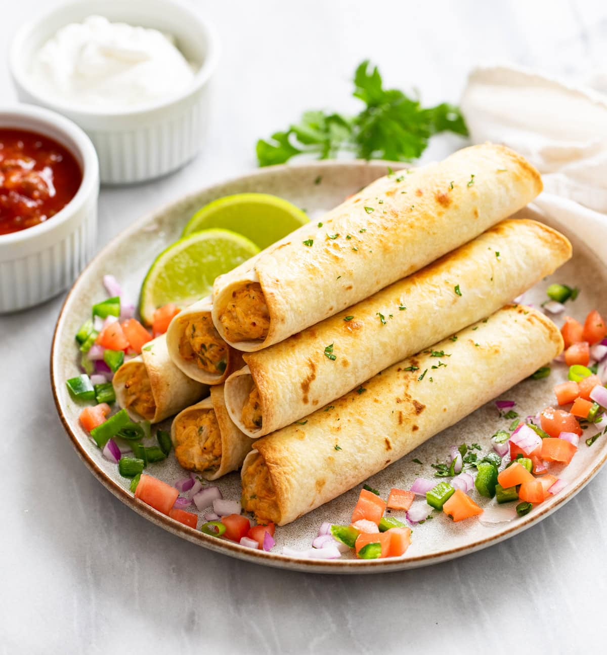 A stack of Chicken Taquitos on a plate surrounded by Mexican toppings with sour cream and salsa in the background.