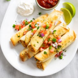 Chicken Taquitos piled up on a white serving platter with diced tomatoes, onions, cilantro, and diced jalapenos.