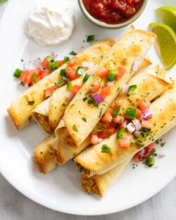 Chicken Taquitos piled up on a white serving platter with diced tomatoes, onions, cilantro, and diced jalapenos.