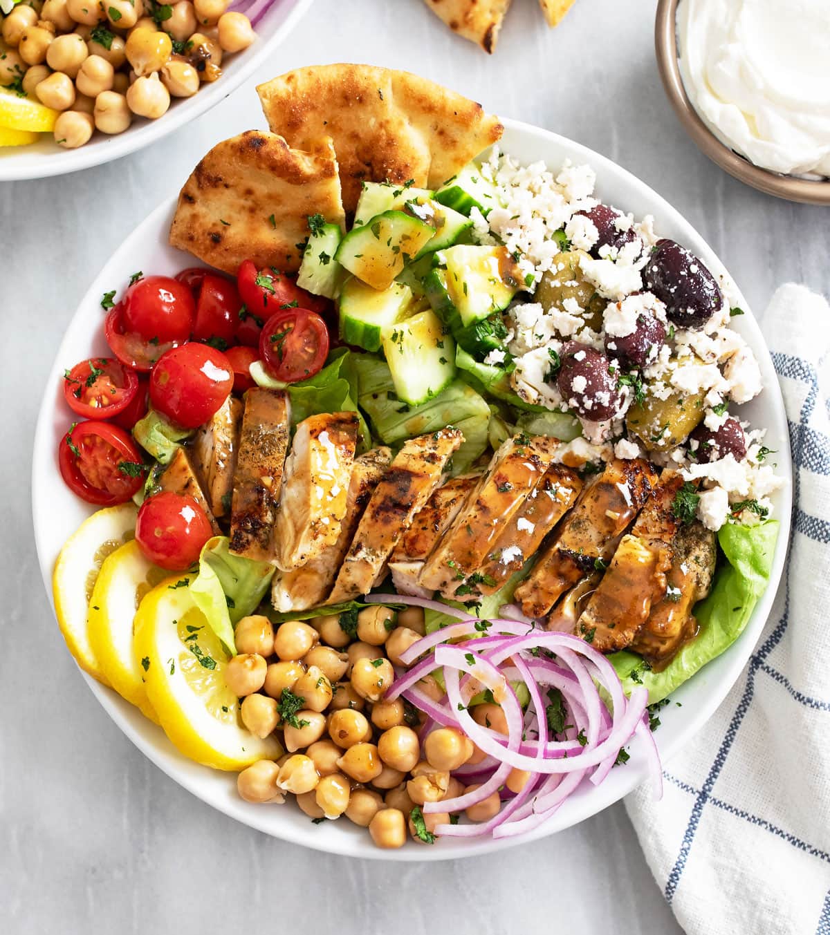 Overhead view of Greek Chicken Bowls in a white bowl with vegetables, olives, feta, and chickpeas.
