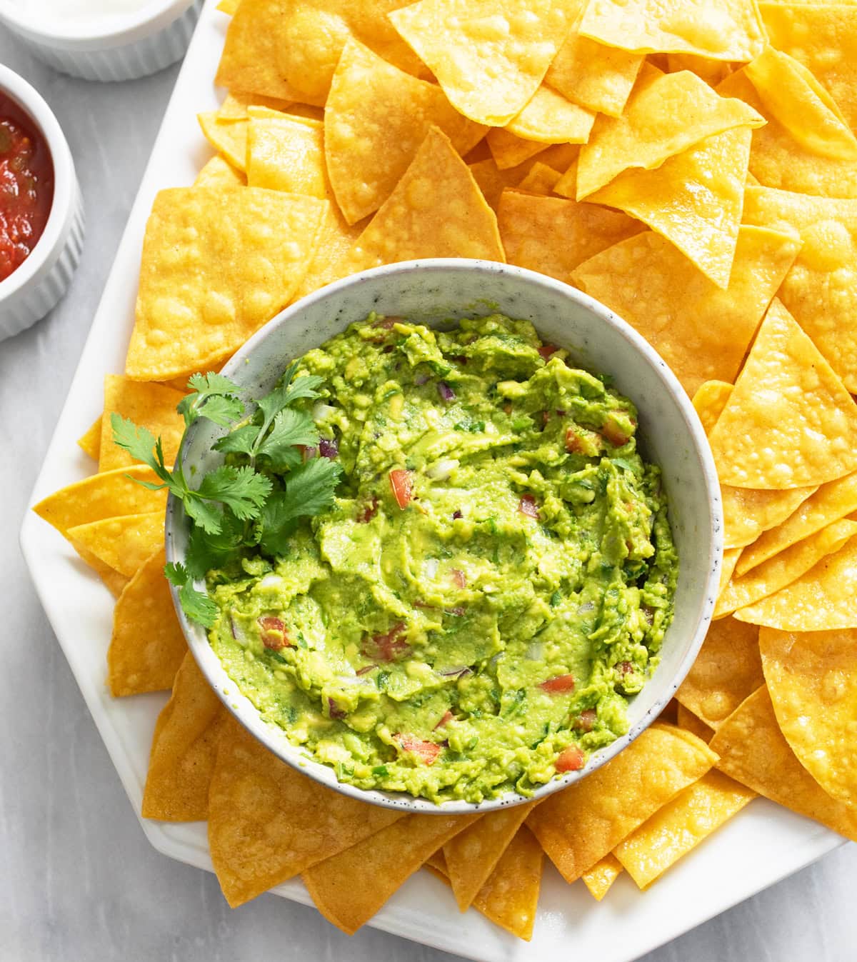 A bowl of Guacamole on a platter with Tortilla Chips and cilantro on the side.