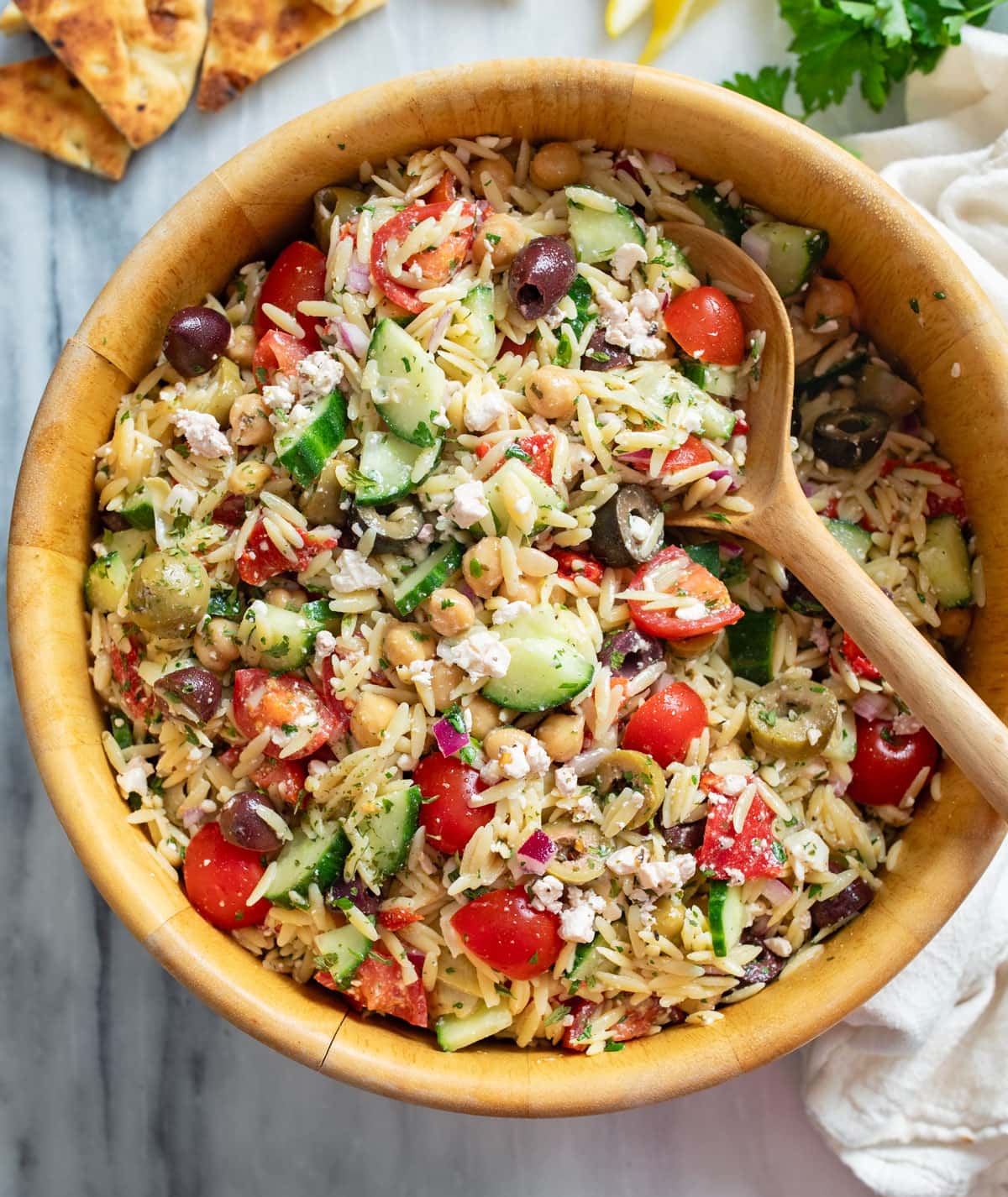 Orzo Salad in a wooden bowl with Mediterranean ingredients and a wooden spoon on the side.