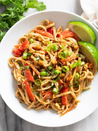 Peanut Butter Noodles in a white bowl with slices of lime on the side and green onions on top.