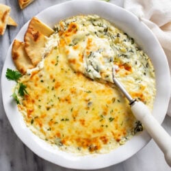 Spinach and Artichoke Dip in a white serving bowl with slices of pita bread on one side and a serving spoon on the other.