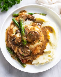 Chicken Madeira next to creamy mashed potatoes on a white plate with mushroom sauce and asparagus.