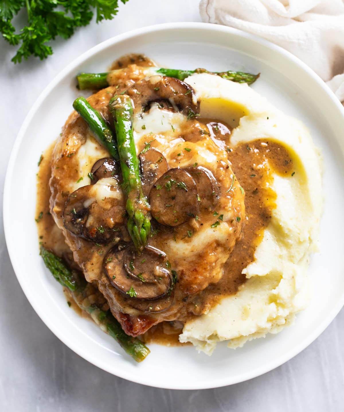 Chicken Madeira next to creamy mashed potatoes on a white plate with mushroom sauce and asparagus.