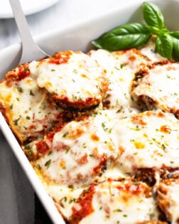 Eggplant Parmesan in a white casserole dish with a spatula scooping it up and fresh basil on the side.