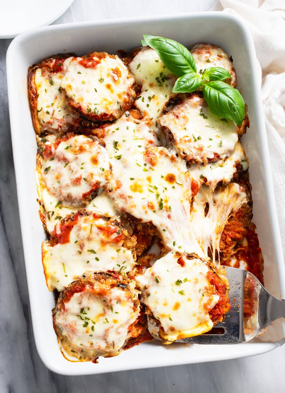 Overhead view of Eggplant Parmesan in a white casserole dish with a spatula scooping it up and fresh basil on the side.