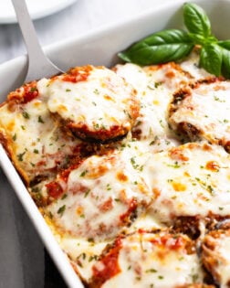 Eggplant Parmesan in a white casserole dish with a spatula scooping it up and fresh basil on the side.