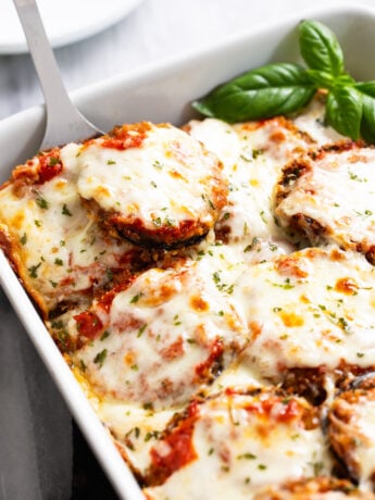 Eggplant Parmesan in a white casserole dish with a spatula scooping it up and fresh basil on the side.
