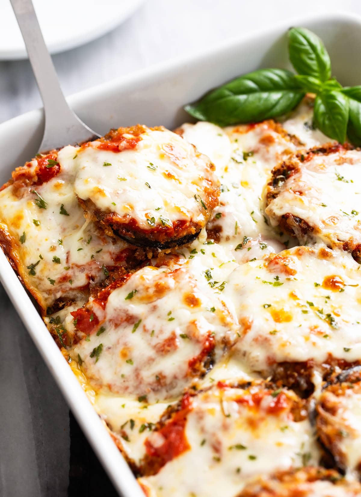 Eggplant Parmesan in a white casserole dish with a spatula scooping it up and fresh basil on the side.