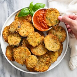 Sliced of Fried Eggplant on a white plate with a ramekin of marinara sauce and fresh basil on the side.