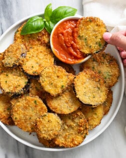 Sliced of Fried Eggplant on a white plate with a ramekin of marinara sauce and fresh basil on the side.