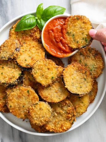 Sliced of Fried Eggplant on a white plate with a ramekin of marinara sauce and fresh basil on the side.