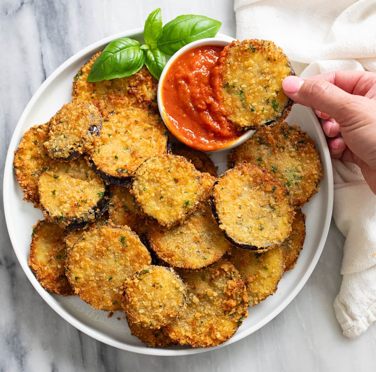 Sliced of Fried Eggplant on a white plate with a ramekin of marinara sauce and fresh basil on the side.