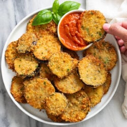 Fried Eggplant on a white plate with a hand dipping one into marinara sauce with fresh basil on the side.