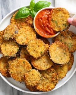Fried Eggplant on a white plate with a hand dipping one into marinara sauce with fresh basil on the side.