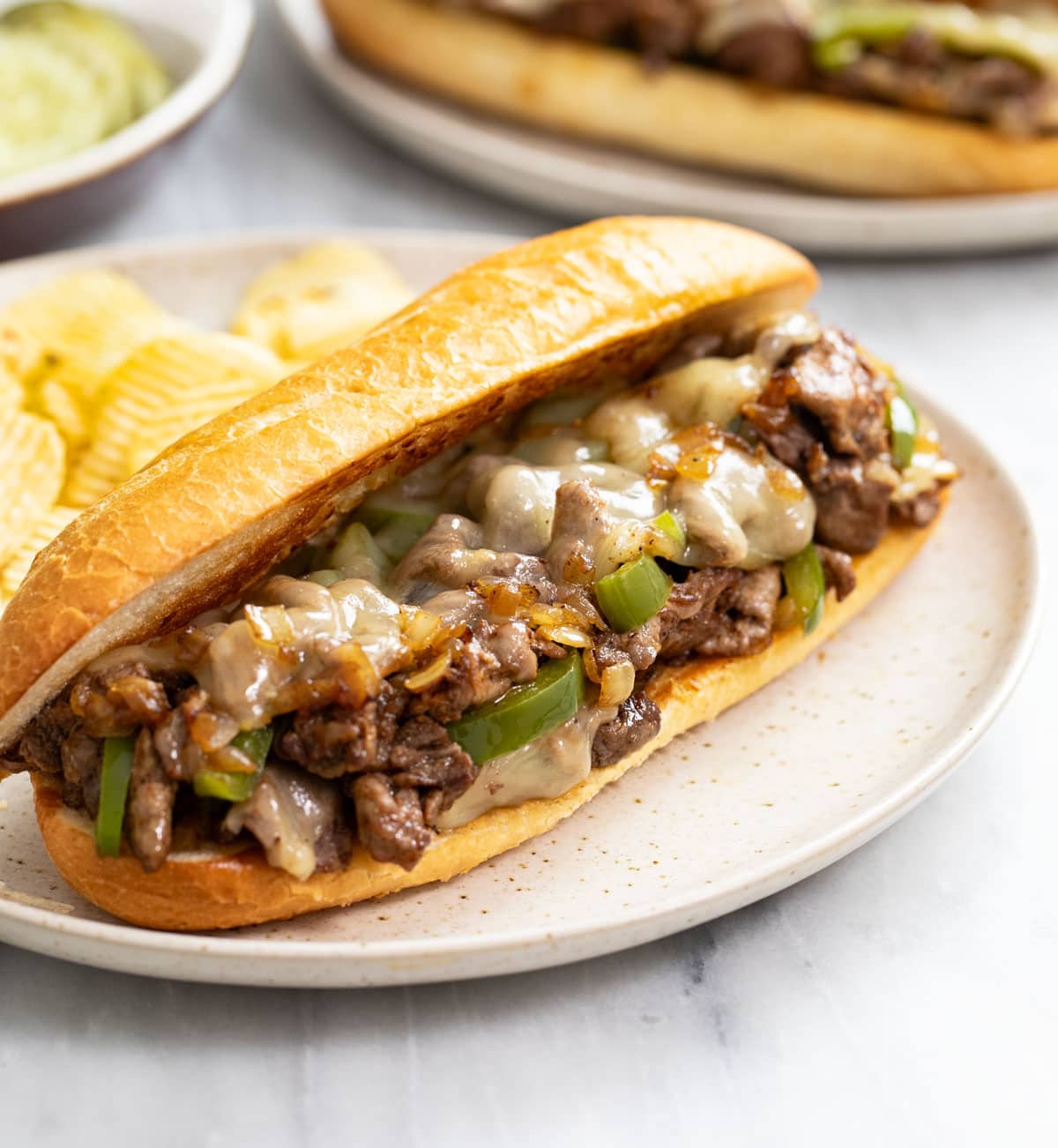 A Philly Cheesesteak Sandwich on a plate with potato chips in the back.