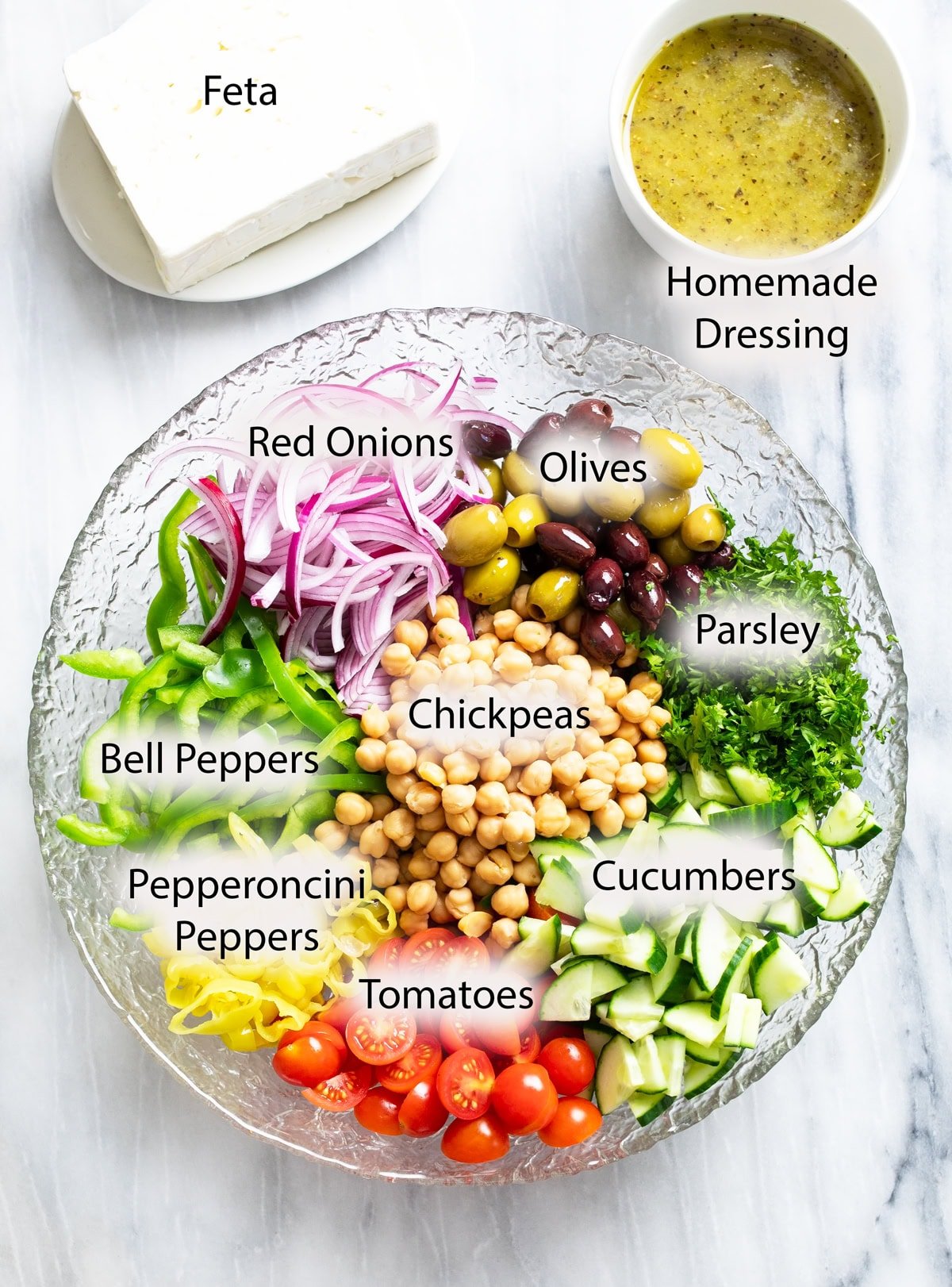 Ingredients for making Greek Salad in a bowl and on a white surface with labels.