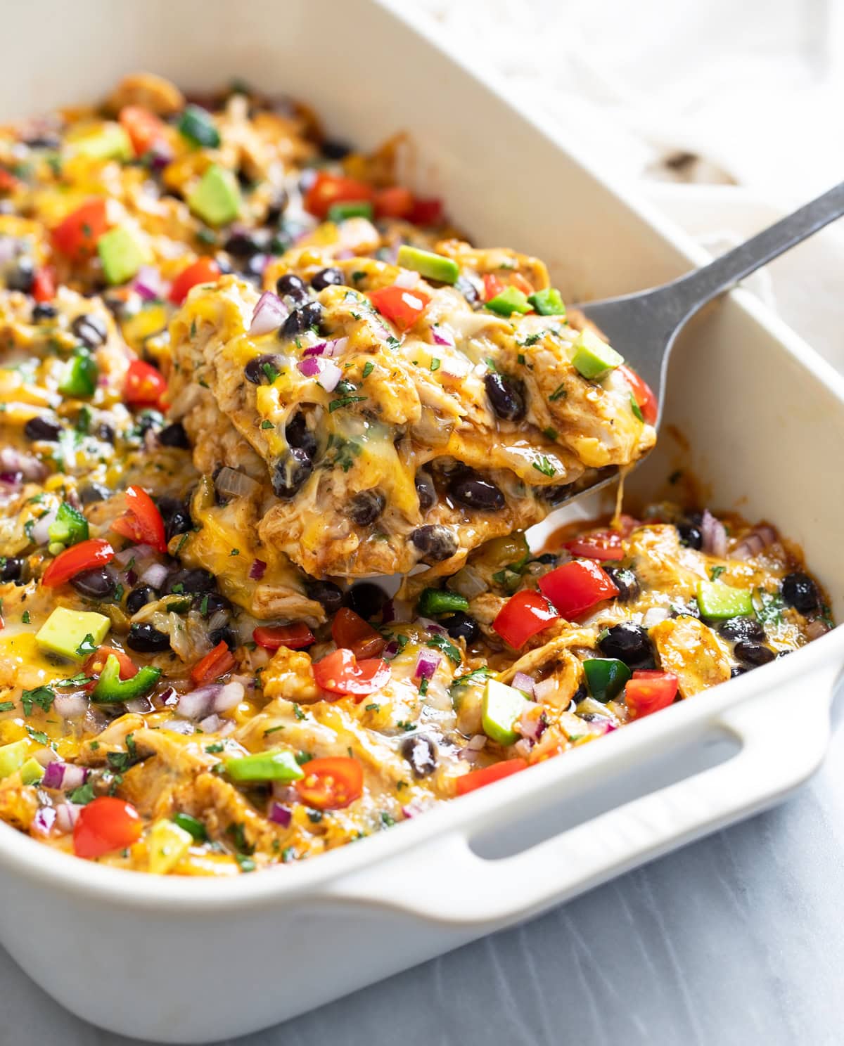 Side view of a spatula lifting out a slice of Chicken Enchilada Casserole from a white baking dish.