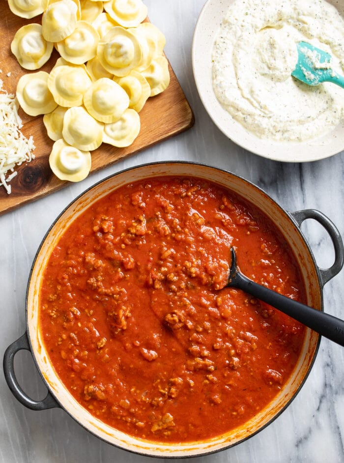 A pot of meat sauce next to a bowl of herb ricotta and a board with tortellini and mozzarella.