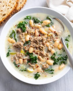 Sausage and White Bean Soup with kale in a white bowl with a spoon on the side.