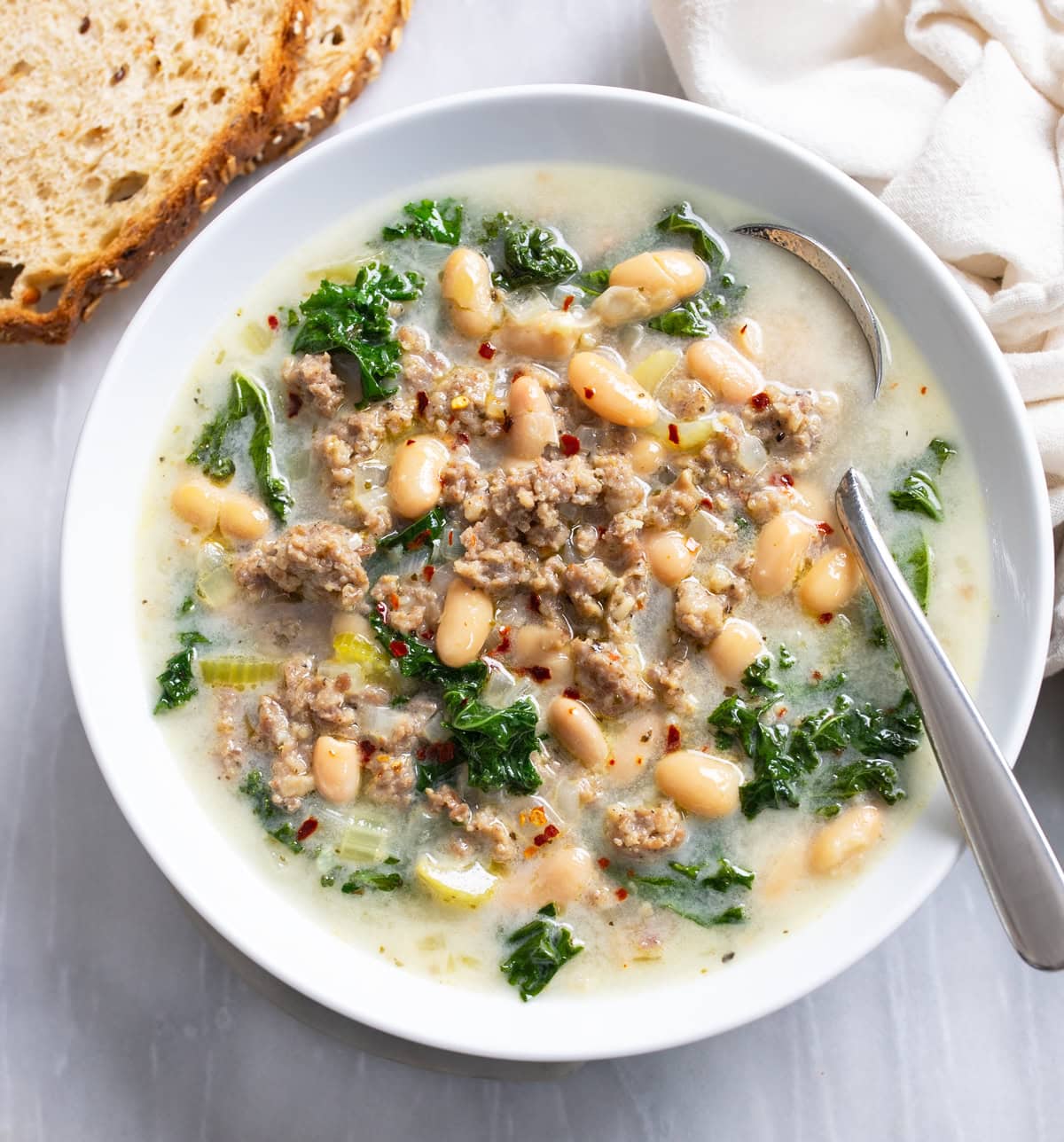 Sausage and White Bean Soup with kale in a white bowl with a spoon on the side.