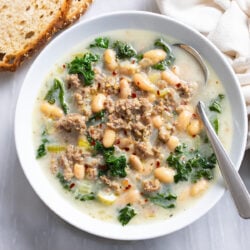 Sausage and White Bean Soup with kale in a white bowl with a spoon on the side.
