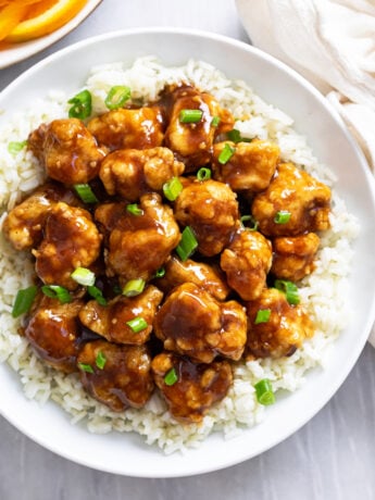 Glossy Orange Chicken on a white plate with rice and green onions.