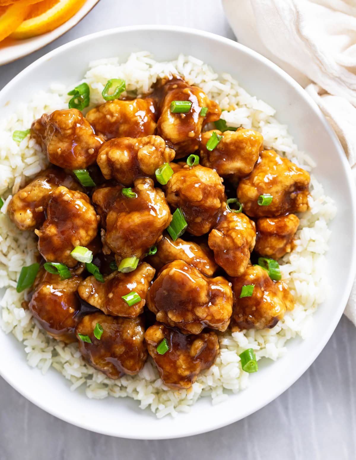 Glossy Orange Chicken on a white plate with rice and green onions.