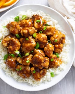 A white plate of Orange Chicken on top of white rice with green onions on top.