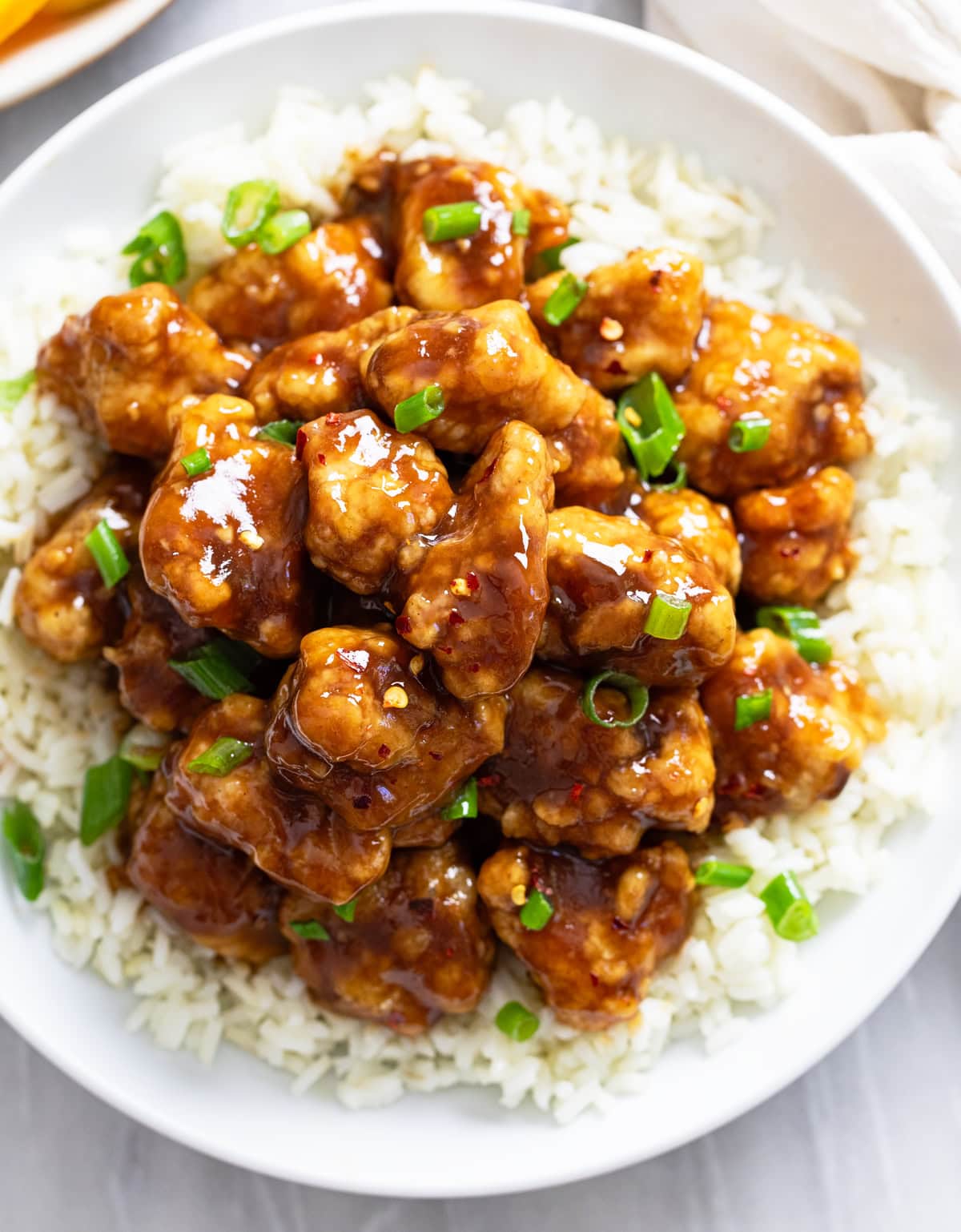 Close up view of a pile of Orange Chicken on white rice with green onions sprinkled on top.
