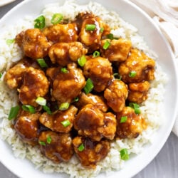 Glossy Orange Chicken over white rice on a white plate with green onions sprinkled on top.