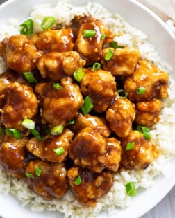 Glossy Orange Chicken over white rice on a white plate with green onions sprinkled on top.