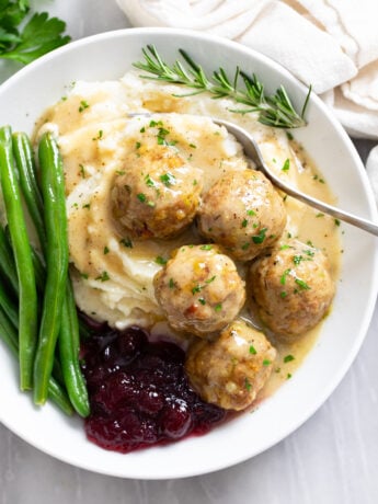 Turkey and Stuffing Meatballs on a white plate with mashed potatoes, gravy, cranberry sauce, and green beans.